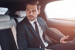 Young and successful. Handsome young man in full suit looking away while sitting in the car photo