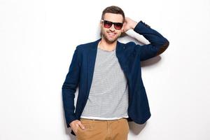 Confident and handsome. Handsome young man in sunglasses holding hand behind head and smiling while standing against white background photo