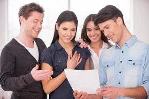 Happy with the result. Group of cheerful young people standing close to each other and smiling while man holding a paper in his hands photo