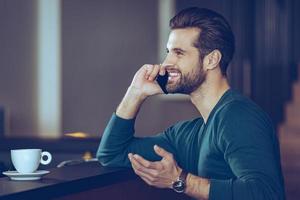 Great phone call. Side view of handsome young man talking on mobile phone with smile while sitting at bar counter photo