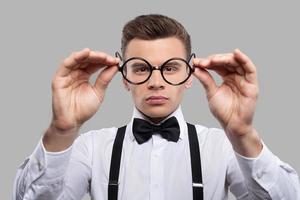 Looking at future. Portrait of thoughtful young man in bow tie and suspenders holding glasses outstretched and looking through it while standing against grey background photo