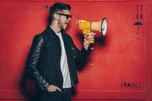 Fashion announcement. Side view of handsome young man in sunglasses holding megaphone and keeping mouth open while standing against red background photo