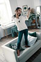 So much fun Full length of attractive young woman wearing headphones and smiling while dancing on sofa at home photo