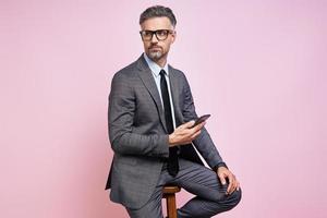Handsome mature man in formalwear holding smart phone while sitting against pink background photo