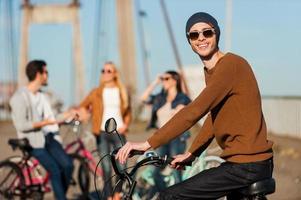 Great day with best friends. Handsome young man on bicycle looking at camera and smiling while his friends talking in the background photo