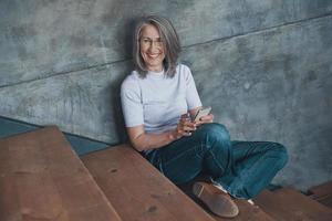 Senior beautiful woman using smart phone and smiling while sitting on the stairs indoors photo