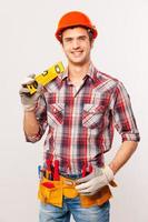 Confident contractor. Handsome young handyman with tool belt carrying work tool on shoulder and smiling while standing against grey background photo