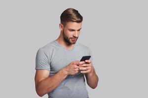 Always online.  Handsome young man holding smart phone and looking at it while standing against grey background photo