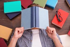 Tired bookworm. Top view of young man covering his face with book while lying on the hardwood floor with many colorful books laying all around him photo