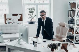 May I help you Handsome young man in full suit looking at camera with serious face while leaning on the office desk photo