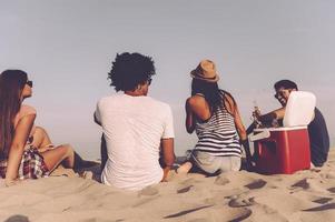 relajante con amigos. vista trasera de cuatro jóvenes alegres que pasan un buen rato juntos mientras se sientan en la playa y beben cerveza foto