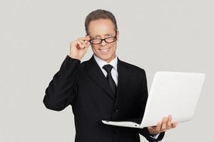 Making your business easy. Cheerful senior man in formalwear holding laptop and adjusting his glasses while standing against grey background photo