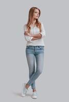 Full length of beautiful young woman in casual wear keeping arms crossed and looking away while standing against grey background photo