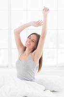 Stretching out before getting up. Cheerful young woman sitting in bed and stretching out while covered with blanket photo