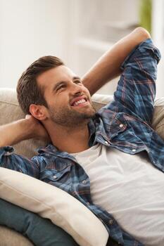 Chill time on sofa. Joyful young man holding hands behind head and smiling while lying on sofa photo