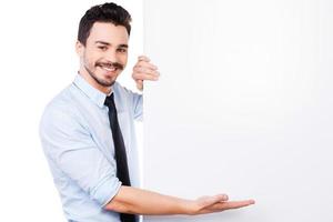 He knows how to sell your product. Handsome young man in shirt and tie leaning at the copy space and pointing it while standing against white background photo