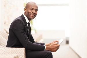 Checking his schedule on the go. Side view of cheerful young African businessman working on digital tablet and smiling at camera photo