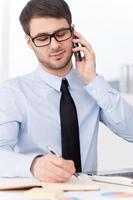 Busy working. Handsome young man in shirt and tie talking on the mobile phone and writing something in note pad while sitting at his working place photo