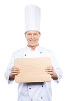 Copy space on his cutting board. Confident mature chef in white uniform holding wooden cutting board and smiling while standing against white background photo