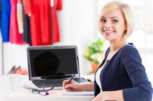 Confident and creative. Beautiful young woman looking over shoulder and smiling while sitting at her working place photo