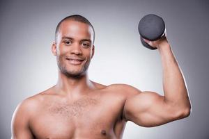 levantamiento de pesas. joven africano sin camisa haciendo ejercicio con pesas y sonriendo mientras está de pie contra el fondo gris foto