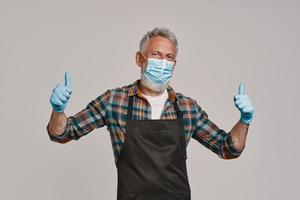 Senior man in apron and protective face mask looking at camera and keeping thumbs up photo