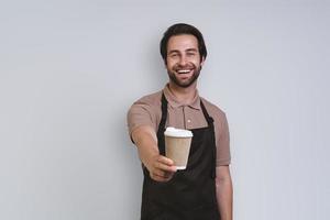 Cheerful young man in apron holding coffee cup and smiling while standing against gray background photo