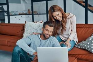 Beautiful young couple using laptop and smiling while spending time at home photo