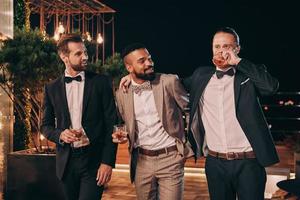 Three handsome men in suits drinking whiskey and communicating while spending time on party photo