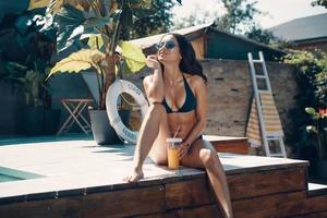 Attractive young woman in bikini drinking cocktail and smiling while sitting by the pool outdoors photo