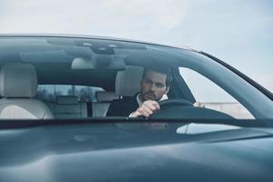 Concentrating on the road. Handsome young man in full suit looking straight while driving a car photo