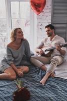 Song for her. Handsome young man playing guitar to his beautiful girlfriend while celebrating Valentine's Day at home photo