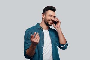 buena plática. encantador joven hablando por teléfono inteligente y sonriendo mientras está de pie contra el fondo gris foto