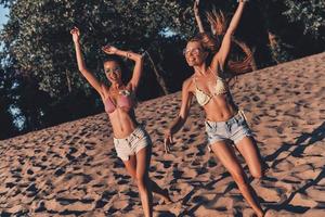 sin preocupaciones dos atractivas mujeres jóvenes en pantalones cortos y trajes de baño sonriendo y gesticulando mientras corren en la playa foto