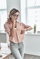 Confident business expert. Beautiful young woman talking on the mobile phone and checking the time while standing in modern office photo