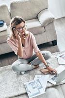 Sharing great ideas. Top view of thoughtful young woman in eyewear working using computer and talking on the phone while flooring at home photo