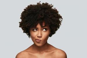 Uncertainty. Confused young African woman looking away and making a face while standing against grey background photo