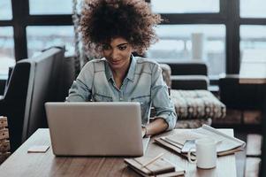 Best job ever. Beautiful young African woman using computer and smiling while sitting at her working place in cafe photo