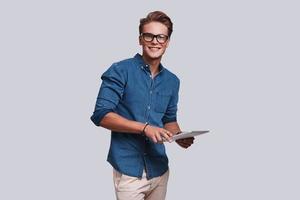 Handsome young man looking at camera and holding digital tablet while standing against grey background photo