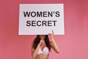 Woman is a secret.  Young woman covering face with poster and gesturing while standing against pink background photo