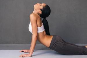 Upward facing dog. Side view of beautiful young African woman in sportswear practicing yoga on exercise mat against grey background photo