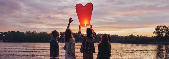 Nothing is better than old friends. Full length of young people in casual wear preparing sky lantern while standing on the pier photo