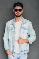 Modern and cool. Confident young man in sunglasses holding hand in pocket and looking at camera while standing against grey background photo