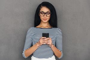 Quick message to friend. Attractive young African woman holding smart phone and looking at it while standing against grey background photo