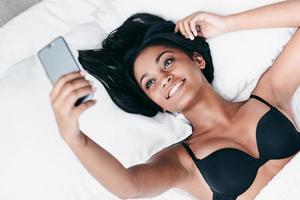 Selfie in the morning. Top view of beautiful young African woman in black lingerie lying in bed and making selfie photo