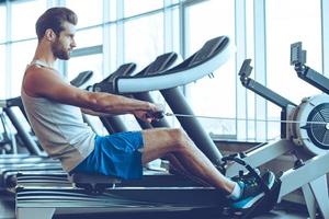 Rowing with power. Side view of young man in sportswear doing rowing in front of window at gym photo