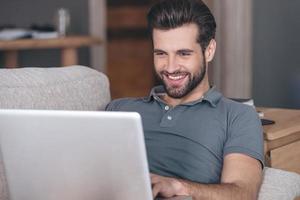 Cheerful and handsome blogger. Cheerful handsome young man using his laptop with smile while sitting on the couch at home photo