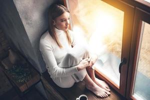 When it is rainy outside. Top view of beautiful young woman looking through window while sitting at windowsill at home photo