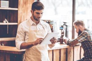 New solutions for his business. Young handsome man in apron using his digital tablet while standing at cafe with customer at background photo
