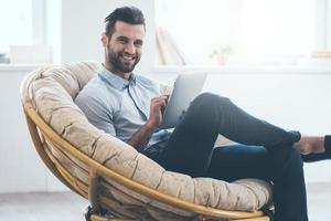 Surfing web from his tablet. Confident young handsome man working on digital tablet and smiling while sitting in big comfortable chair at home photo